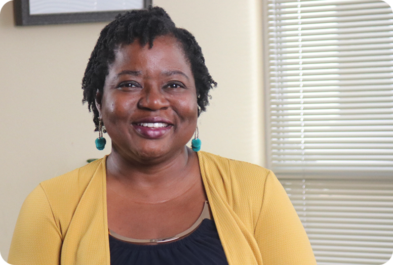 A link to a video. A smiling Black woman named Vernelia with green earrings wearing a yellow cardigan over her black top.